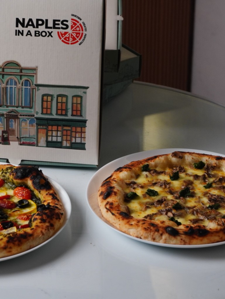 Neapolitan pizzas beside a Naples in a Box takeaway box with storefront illustration